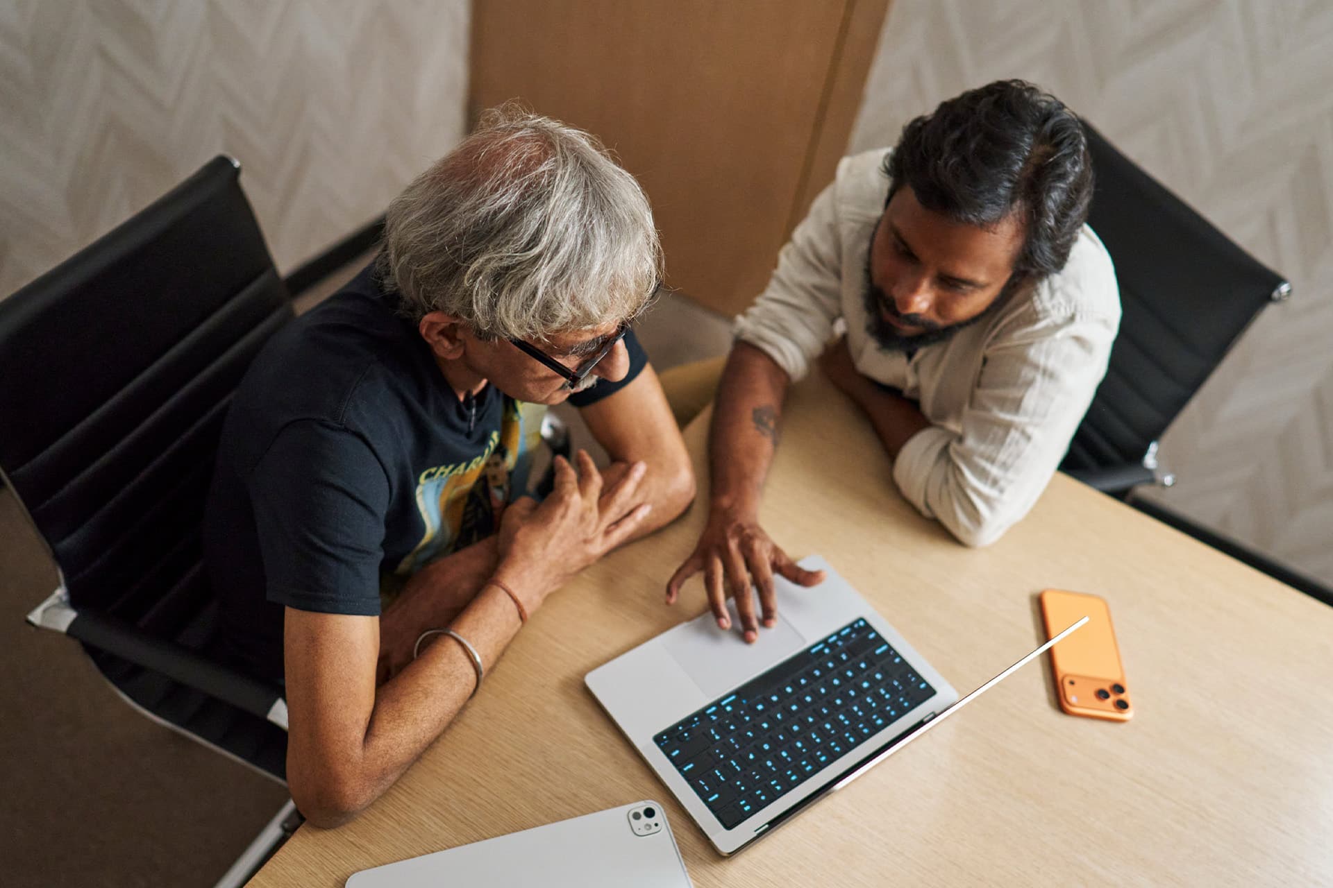 Two people sit at a desk looking at a laptop screen together. One points at the laptop’s keyboard while the other watches attentively. A tablet and a smartphone lie on the table nearby. Filmed on iPhone.