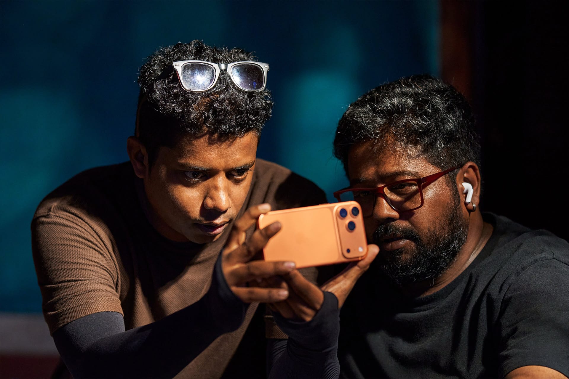 Two men are closely looking at a smartphone, filmed on iPhone. One wears glasses on his head, the other wears glasses and a wireless earbud. Both appear focused, with a blue background behind them.