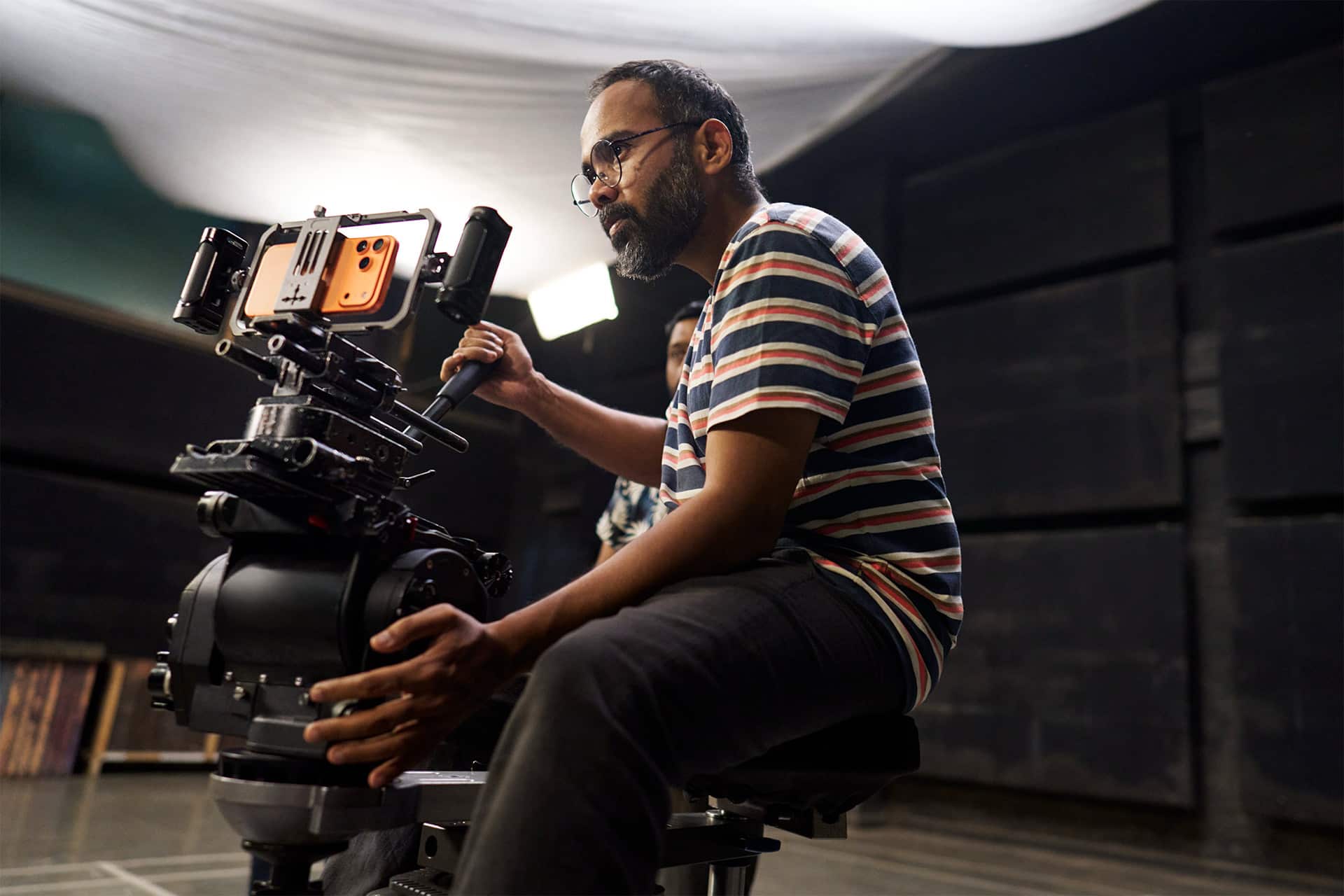 A man wearing glasses and a striped shirt operates a professional video camera on a tripod indoors, focusing intently on his work in a dimly lit studio setting—capturing moments as if they were filmed on iPhone.