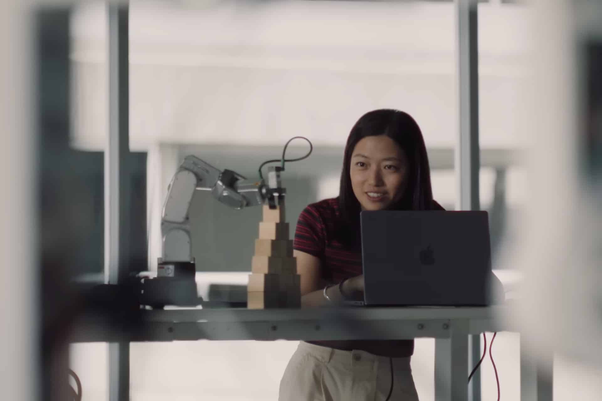 A woman stands at a desk with a laptop and a robotic arm stacking wooden blocks, smiling and looking engaged in her work—an inspiring scene for Mac Students passionate about technology and innovation.