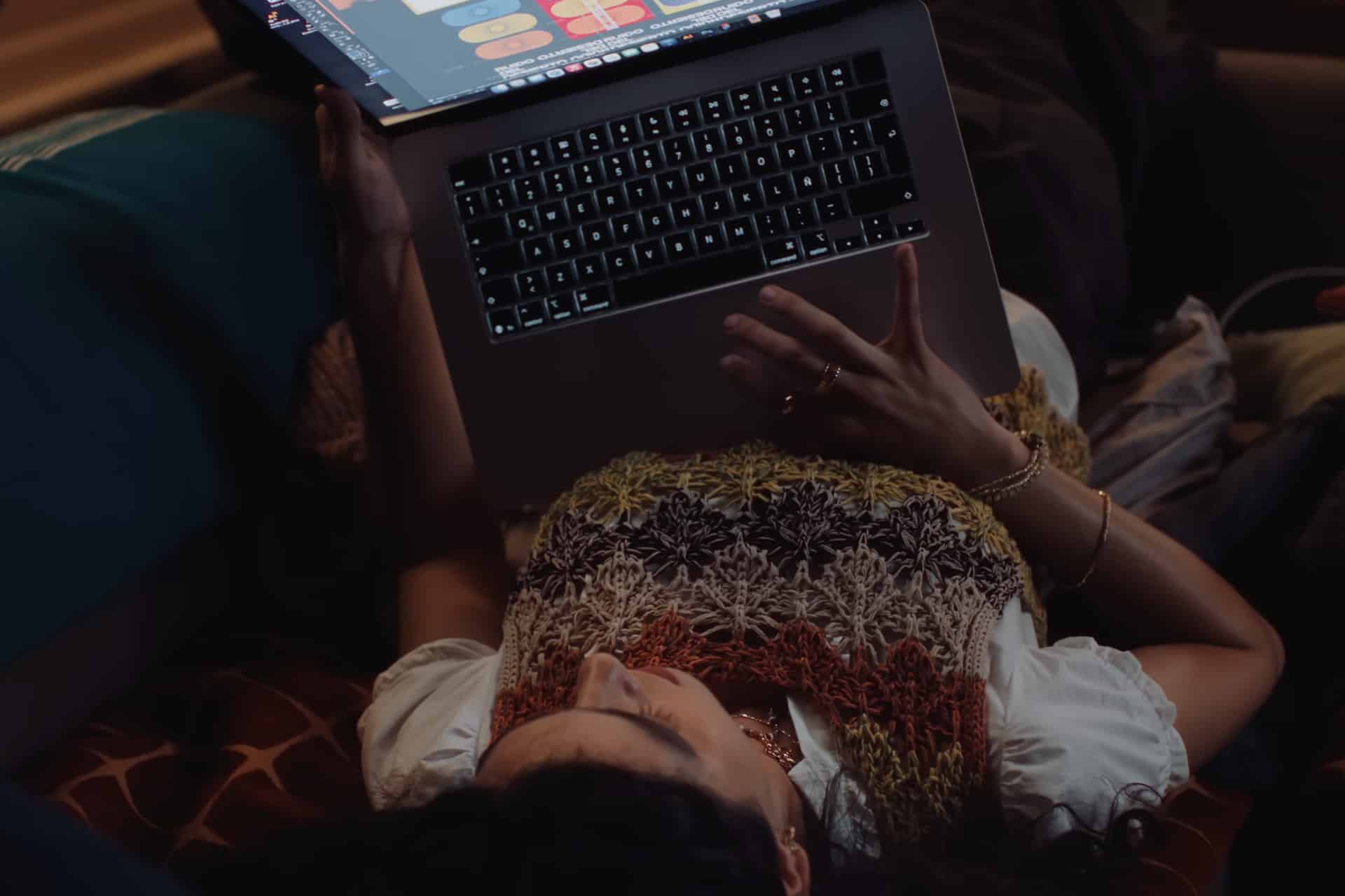 A Mac Student lies down holding a laptop above them, one hand on the keyboard. They wear a colorful crochet vest over a white shirt, and the laptop screen displays a vibrant interface.