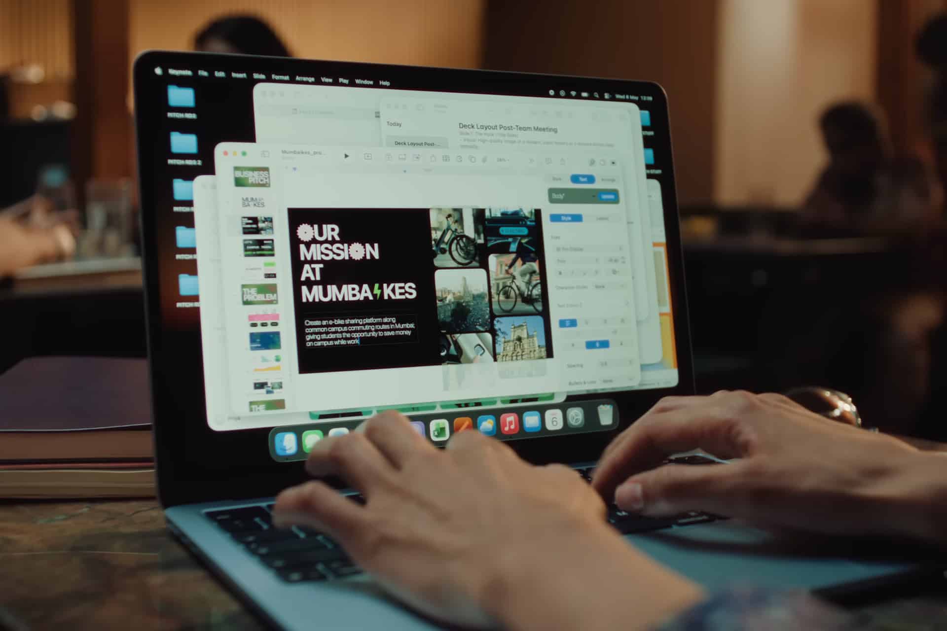 Hands typing on a laptop displaying multiple windows, including a presentation slide that says "OUR MISSION AT MUMBAIKES" and various images—perfect for Mac Students. The background shows blurred people working.