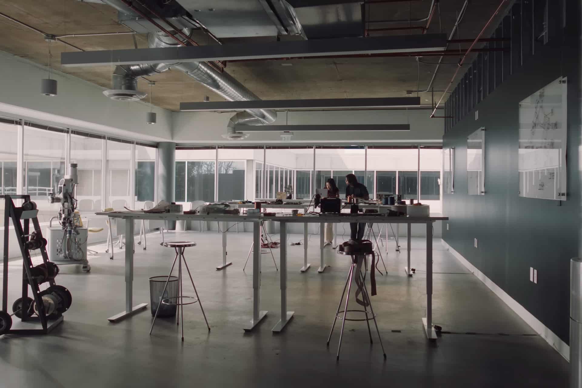 A modern, spacious office with high ceilings, large windows, and exposed ducts. Two Mac Students stand at a tall table covered with papers and models. Robots and stools are visible around the minimalist workspace.