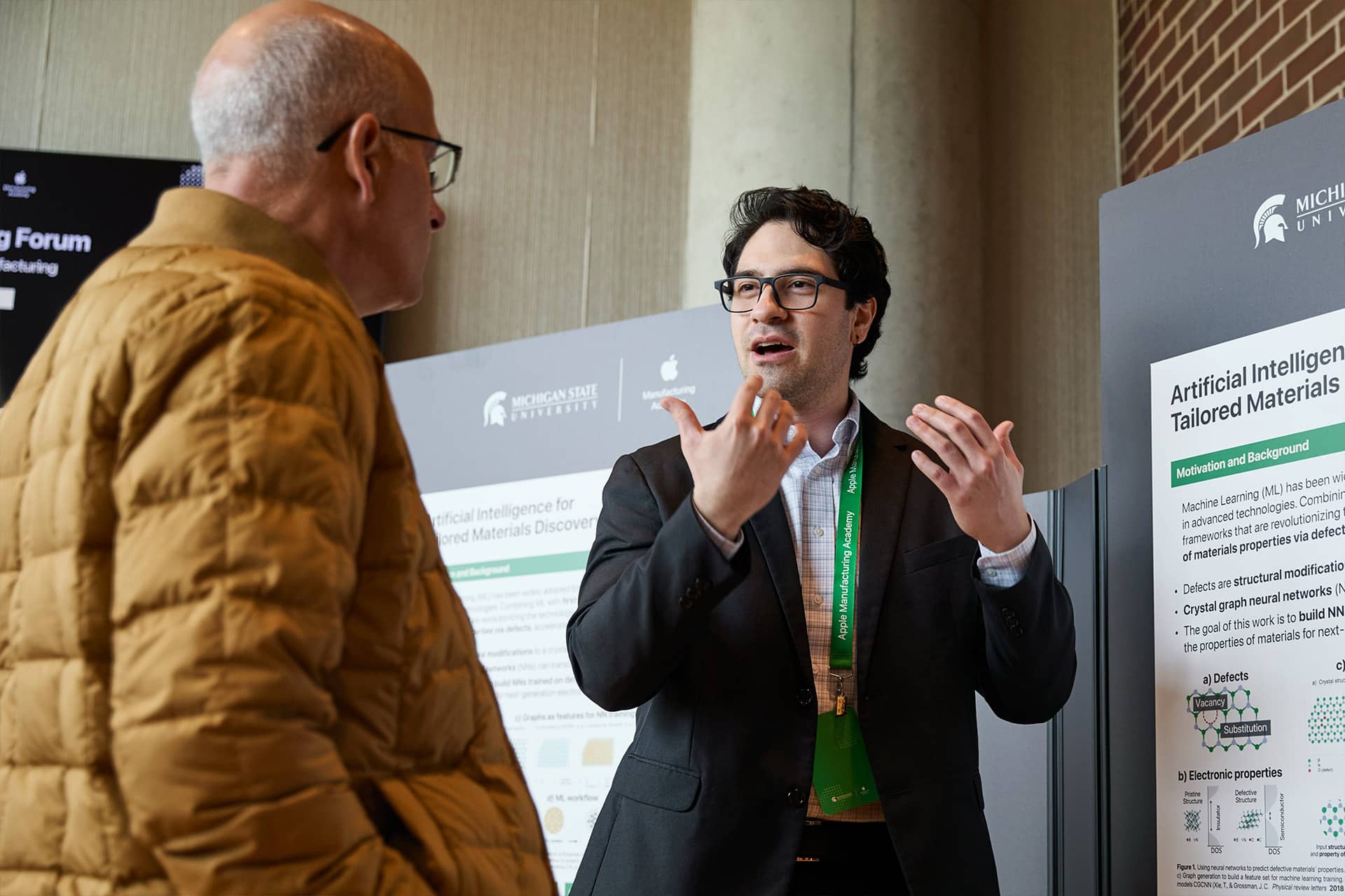 Apple Manufacturing Academy - A man wearing glasses and a suit explains a scientific poster about artificial intelligence and materials discovery to another man in a yellow jacket at a conference.