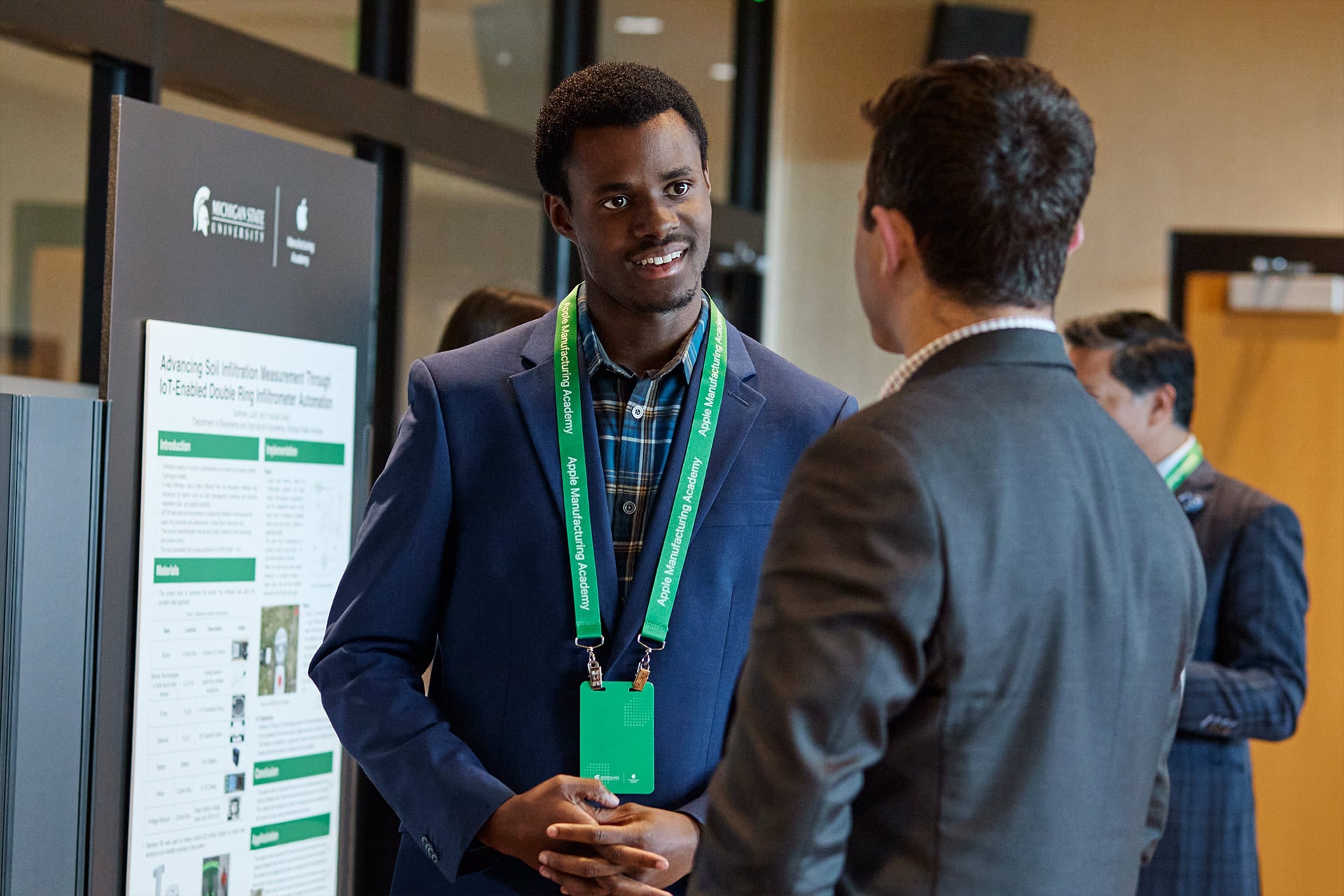 A man in a blue suit and green lanyard speaks with another man in a gray suit at a conference or academic event, standing near a presentation board with Michigan State University branding.