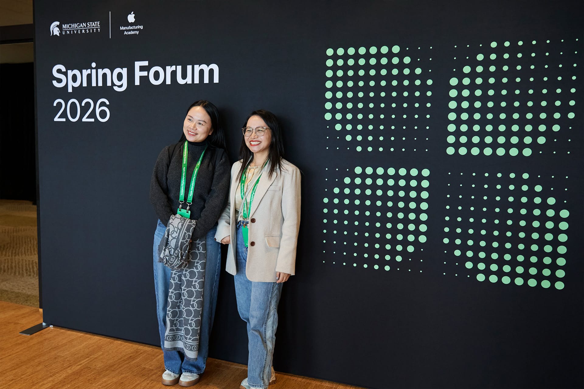 Two women stand smiling in front of a black backdrop that reads “Spring Forum 2026” with Michigan State University and Apple Learning Academy logos. Both wear green lanyards and casual outfits.