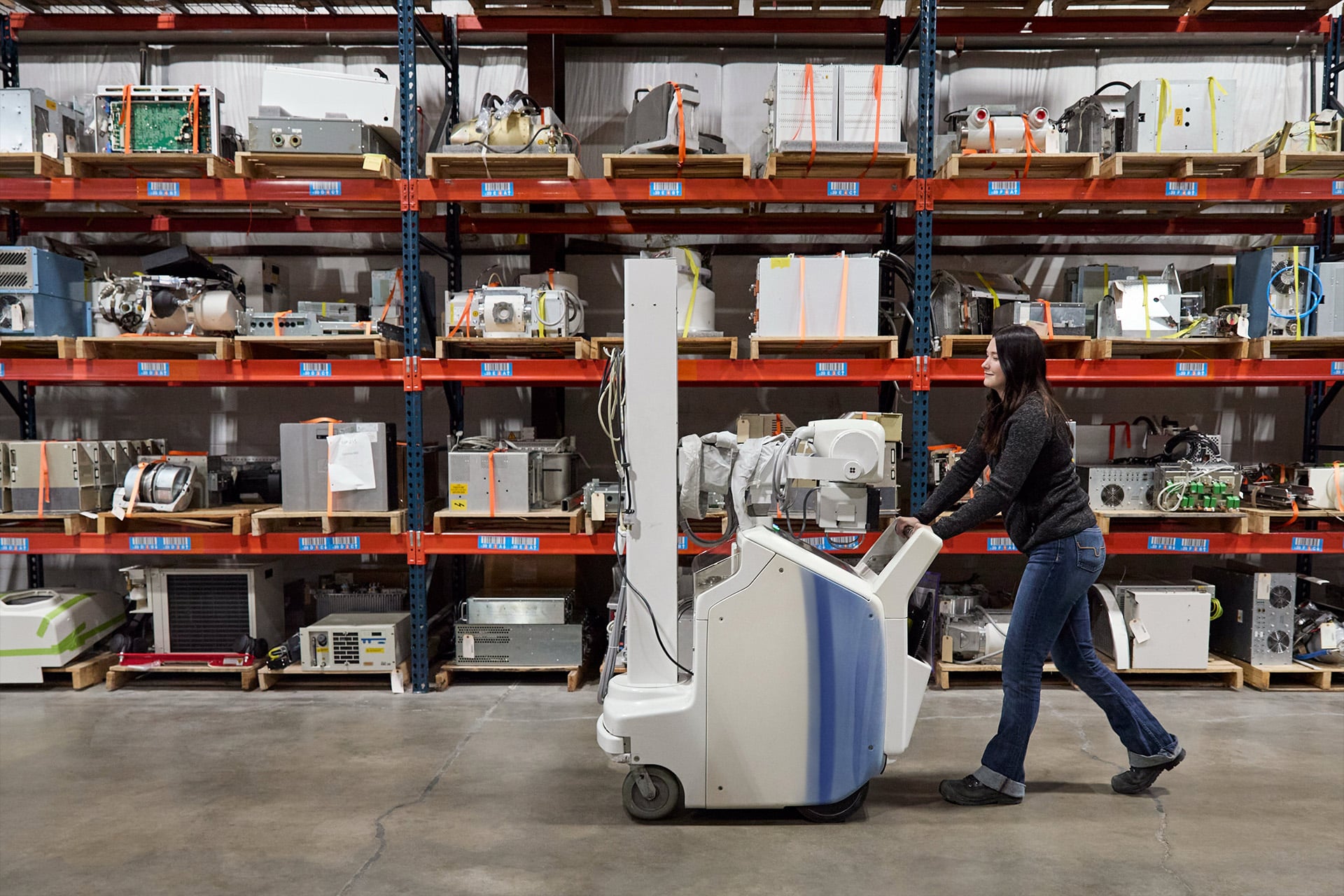 A woman pushes a large medical imaging machine past shelves filled with various electronic and medical devices in a warehouse setting.