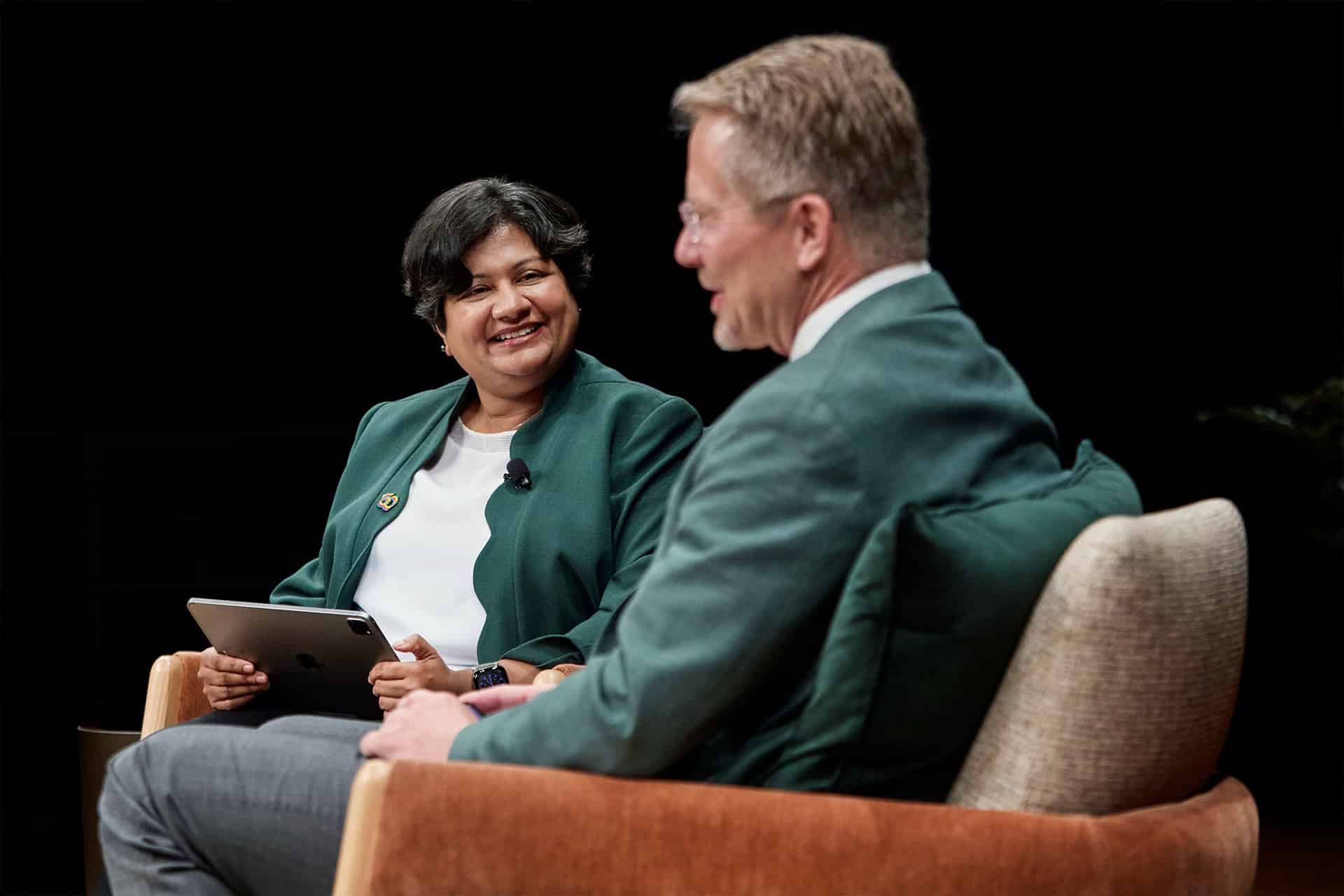 Two people sit on stage in armchairs, engaged in conversation. The woman on the left holds a tablet and smiles at the man on the right. Both wear green jackets and appear to be participating in an event or panel discussion.