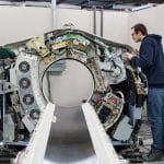 Two people work on the open and exposed interior of a large medical imaging machine, likely a CT scanner, in a bright, industrial setting.