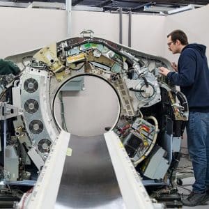 Two people work on the open and exposed interior of a large medical imaging machine, likely a CT scanner, in a bright, industrial setting.
