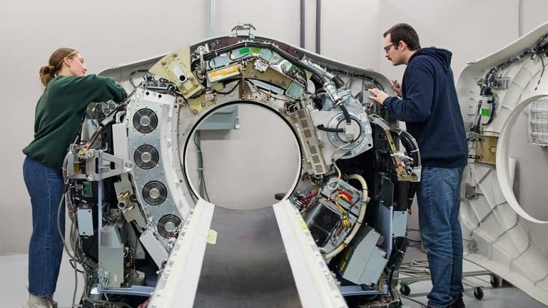 Two people work on the open and exposed interior of a large medical imaging machine, likely a CT scanner, in a bright, industrial setting.