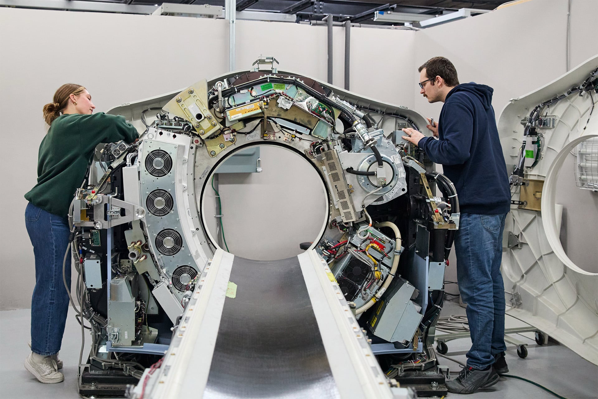 Two people work on the open and exposed interior of a large medical imaging machine, likely a CT scanner, in a bright, industrial setting.