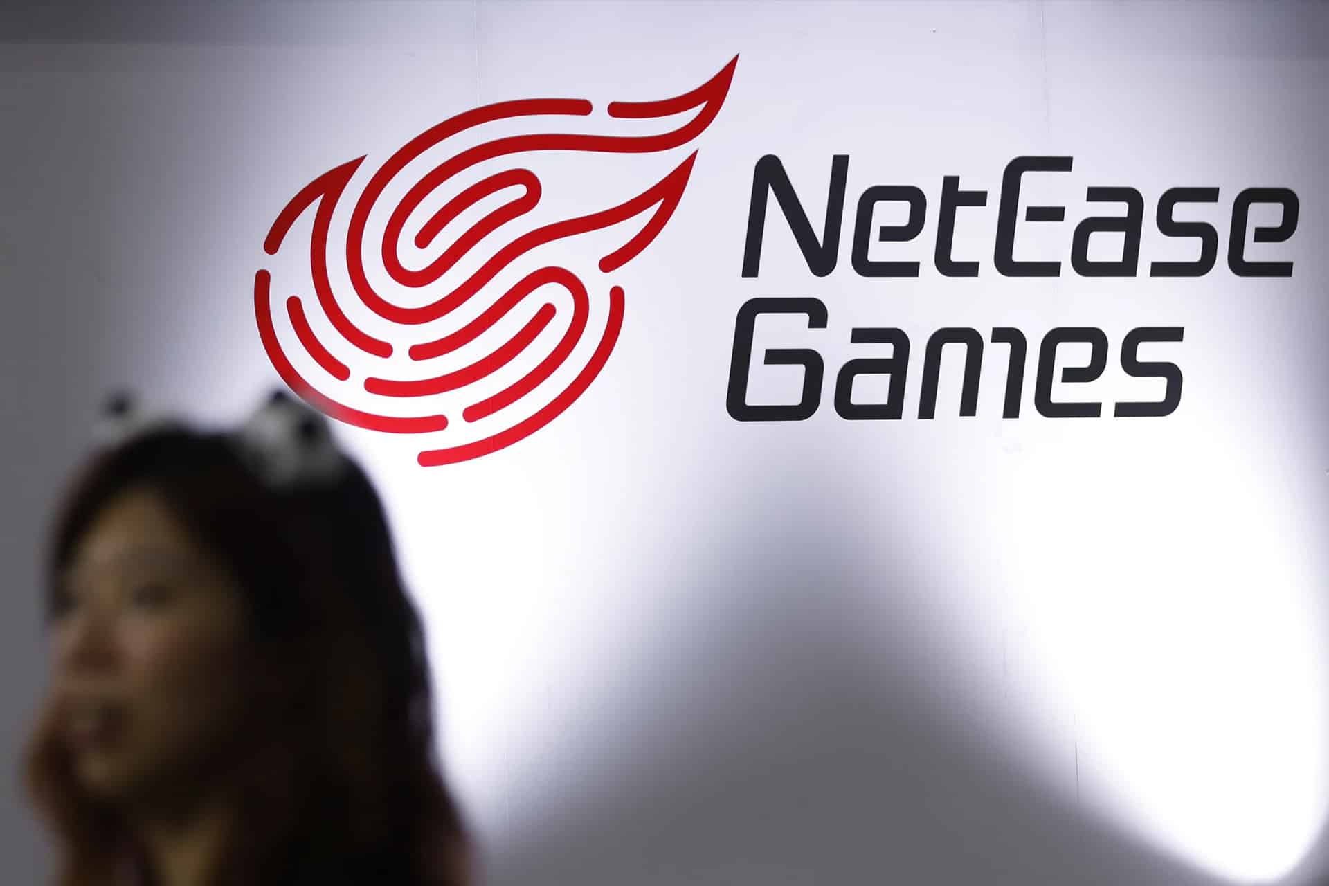 A blurred person stands in front of a white wall with the red NetEase Games logo and black "NetEase Games" text, two brands recognized as major competitors alongside Tencent in the gaming industry.