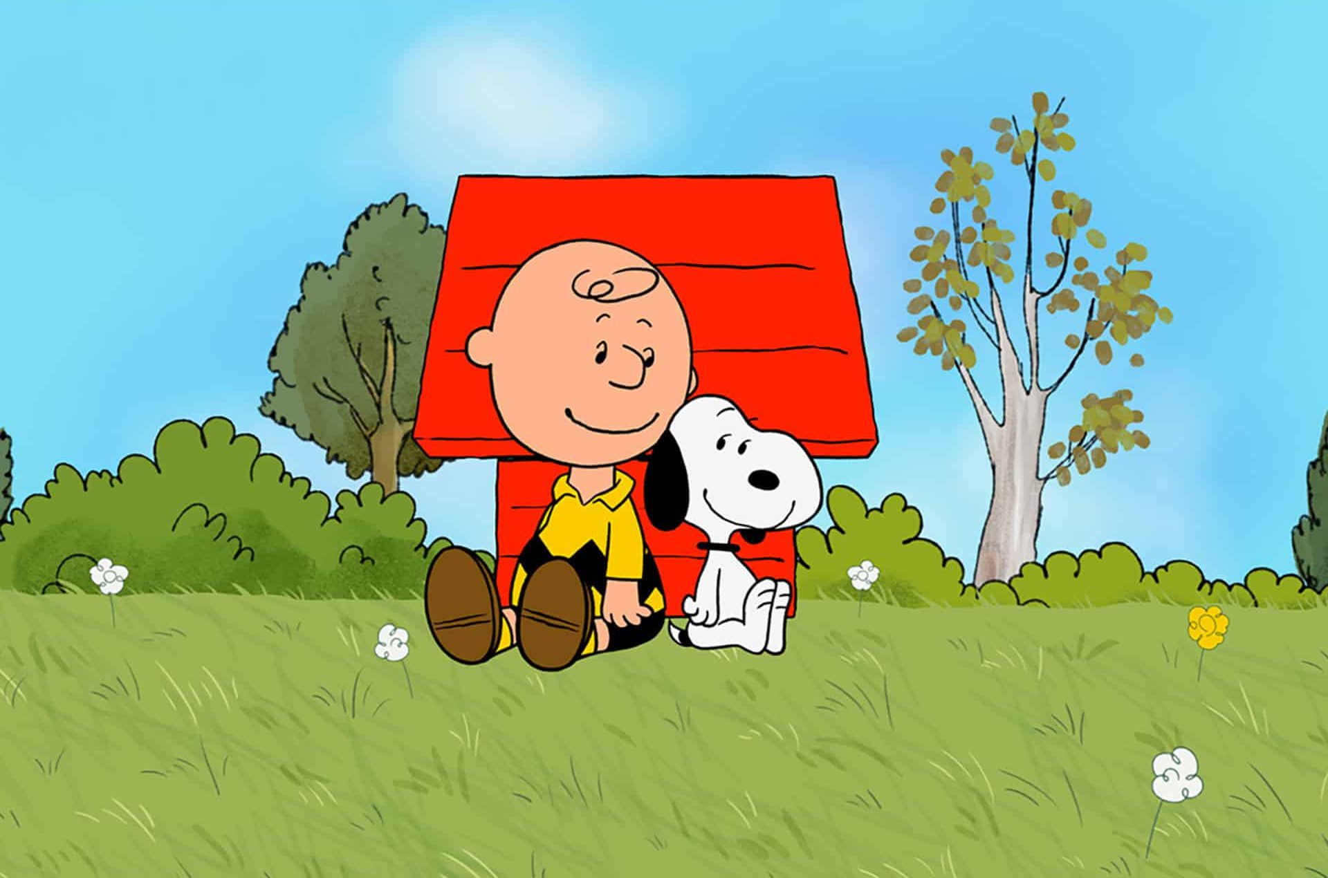 Cartoon boy in a yellow shirt and his white dog sit together on grass before a red doghouse. Green bushes, trees, and a blue sky with clouds complete this Peanuts Apple TV-inspired scene.