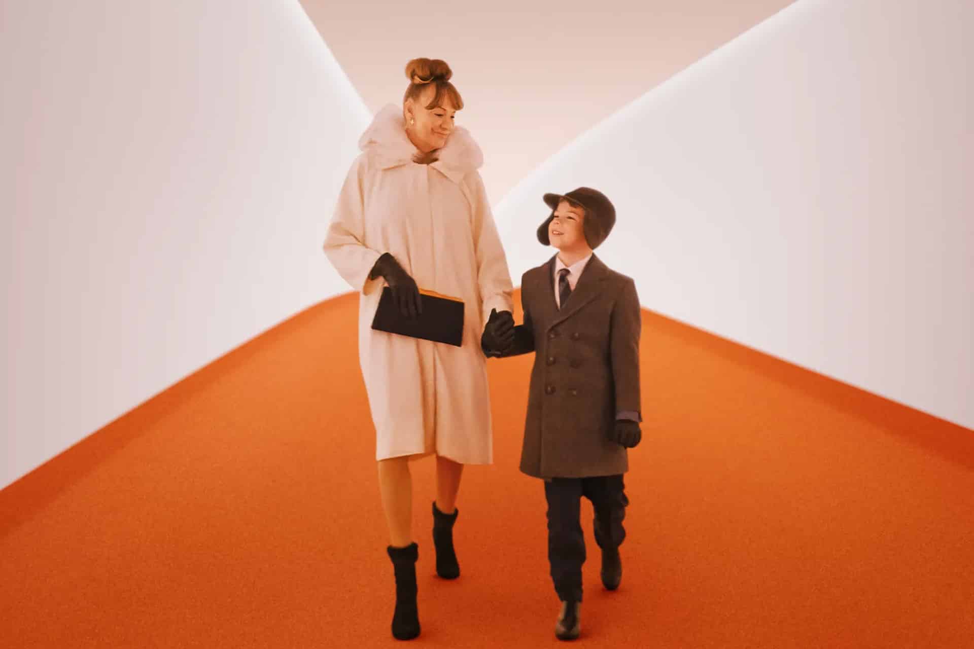 A woman and a young boy, both dressed in stylish coats and winter accessories, walk hand in hand down a bright hallway with orange flooring and white walls, sharing smiles like travelers boarding the Propeller One-Way Night Coach.