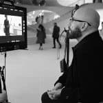 A bald man with glasses sits and watches a director’s monitor on a film set. The monitor displays a scene, reminiscent of Propeller One-Way Night Coach, filmed in a modern building as crew members stand nearby and people walk in the background.