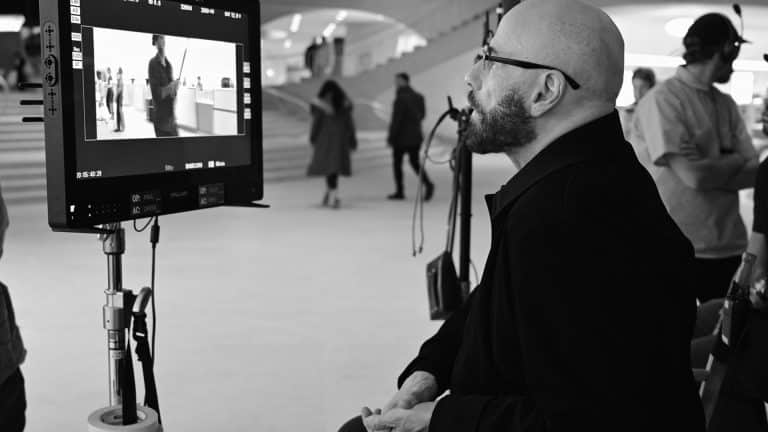 A bald man with glasses sits and watches a director’s monitor on a film set. The monitor displays a scene, reminiscent of Propeller One-Way Night Coach, filmed in a modern building as crew members stand nearby and people walk in the background.
