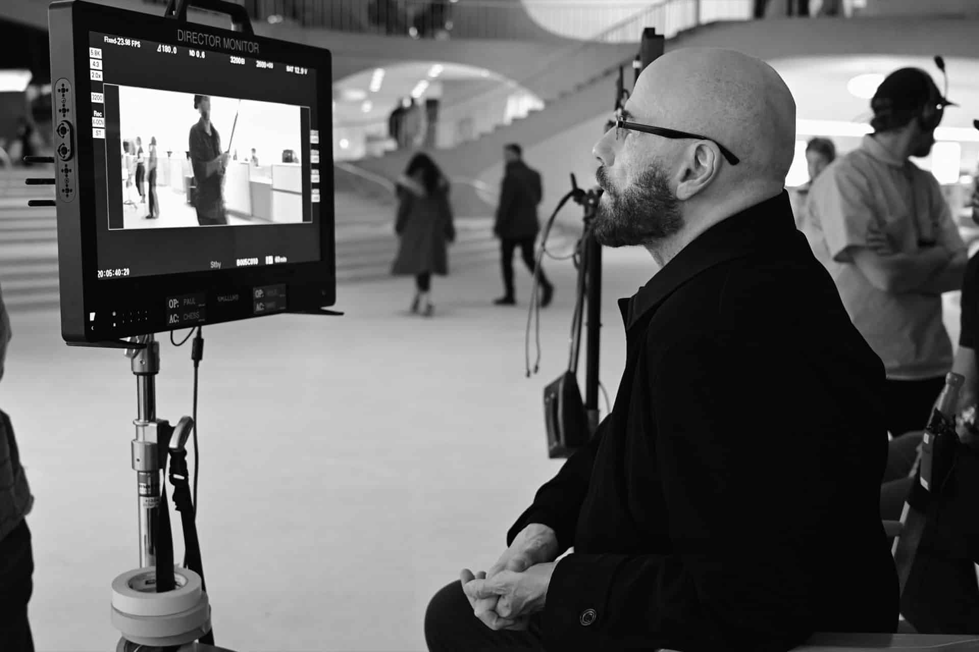A bald man with glasses sits and watches a director’s monitor on a film set. The monitor displays a scene, reminiscent of Propeller One-Way Night Coach, filmed in a modern building as crew members stand nearby and people walk in the background.