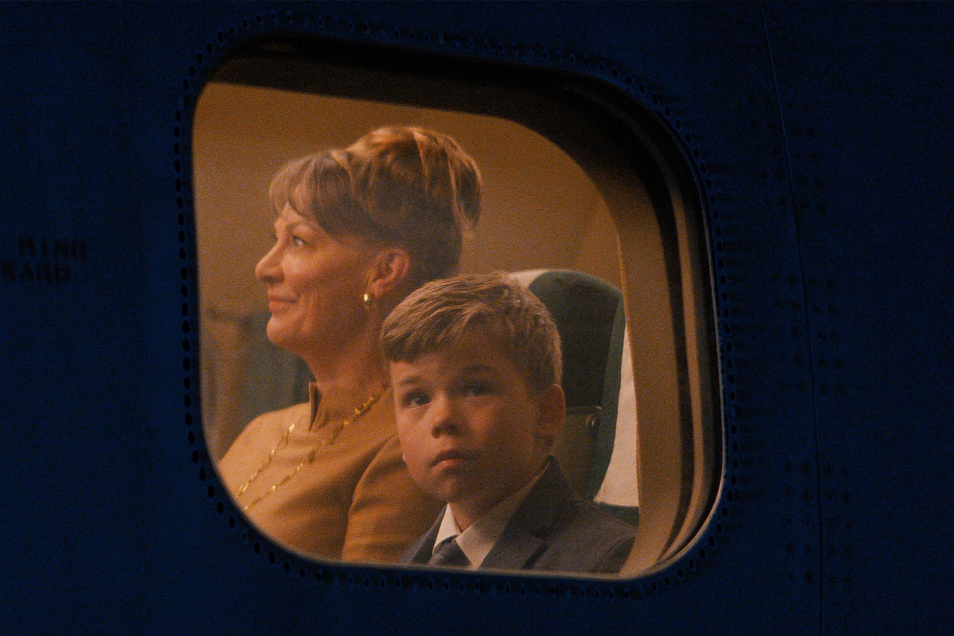 A woman and a boy sit side by side on a Propeller One-Way Night Coach, gazing out the airplane window. The woman smiles gently while the boy looks ahead with a serious expression, both framed against the night sky outside.