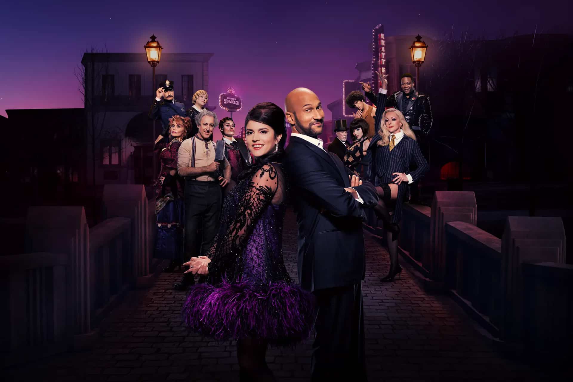 A man and woman in formal, vintage-style attire stand back to back on a cobblestone bridge at dusk, as if part of a Schmigadoon revival, surrounded by colorfully dressed people posing dramatically under old-fashioned street lamps.