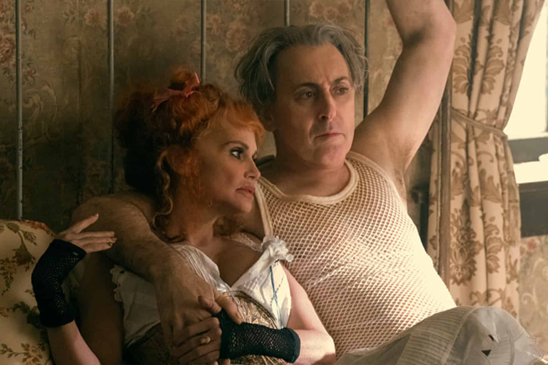 A woman with curly red hair and a man with gray hair sit closely together on a couch, echoing the vibrant style of a Schmigadoon revival. He wears a white mesh undershirt and has his arm around her as she dons a corset and black fingerless gloves.