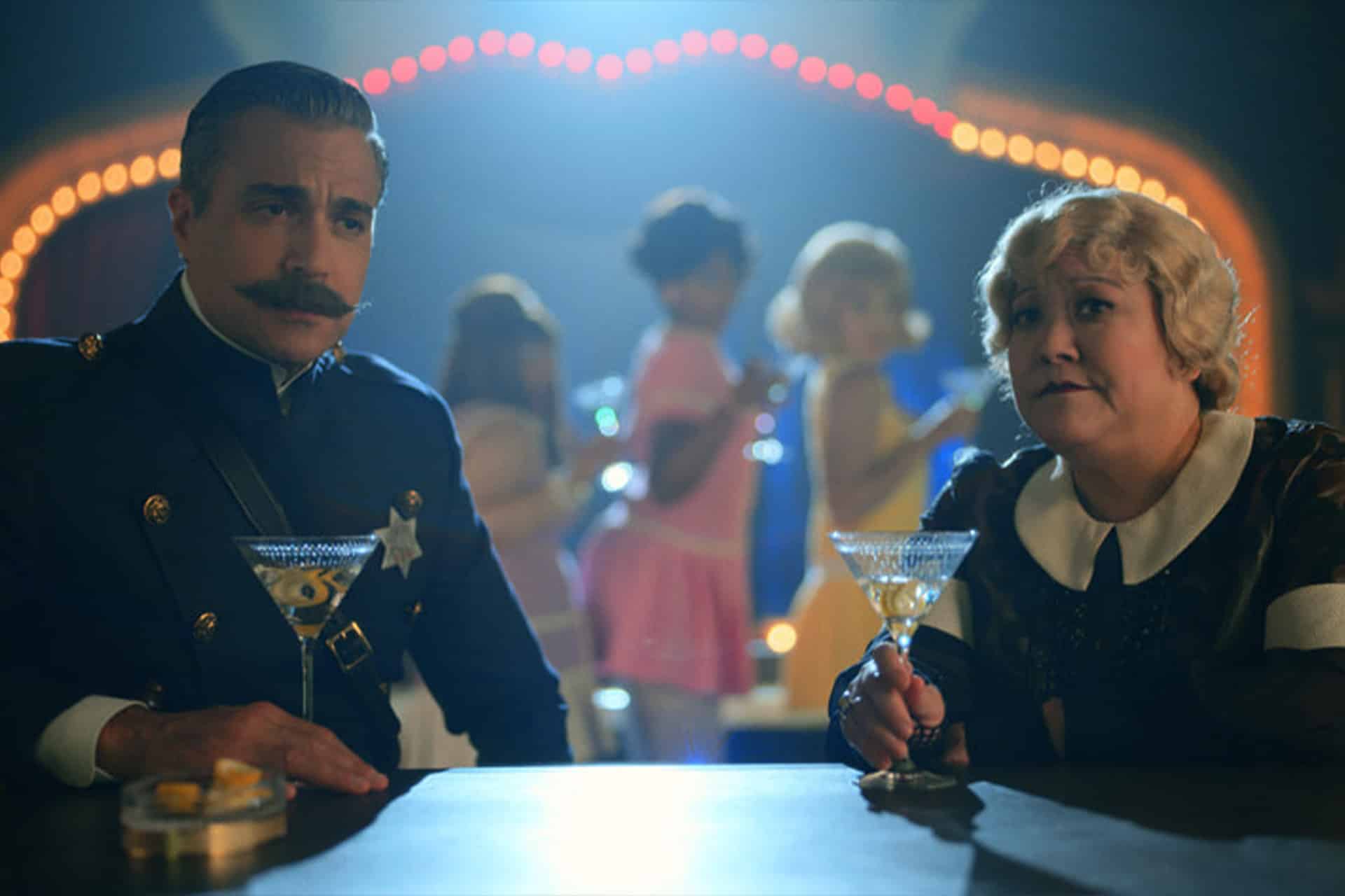 A man in a police uniform and a woman with short blonde hair sit at a bar holding cocktails, as people dance under colorful lights—a lively scene reminiscent of a Schmigadoon revival.