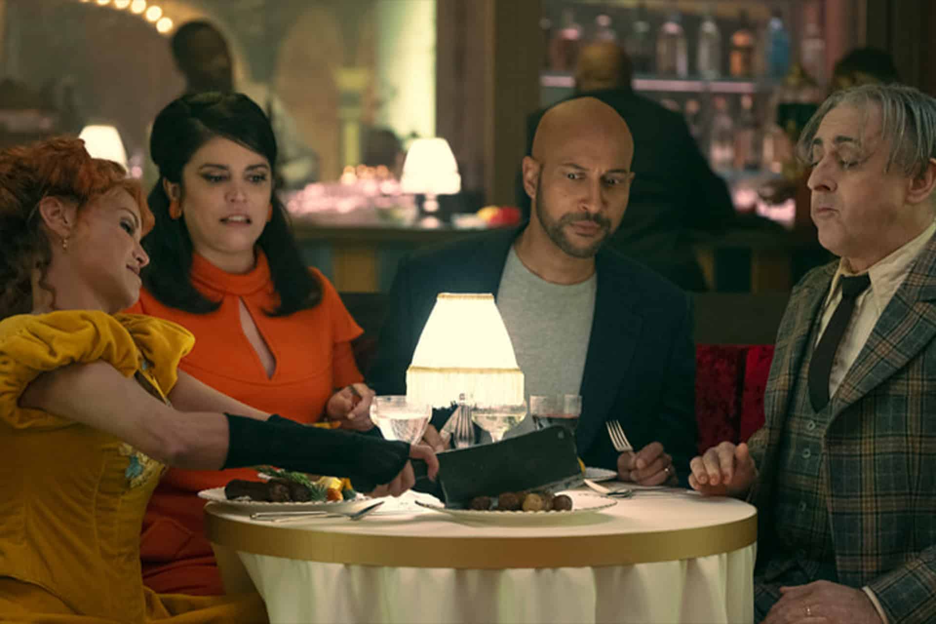 Four people sit around a small, elegantly set dinner table in a dimly lit restaurant. One woman in a bright yellow dress, reminiscent of a Schmigadoon revival, dramatically cuts her food while the others look on with mixed expressions.