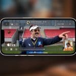 A smartphone displays a scene from the series "Ted Lasso" on Apple TV+, as a man with a mustache points and smiles. Thanks to Apple TV SharePlay, a small video call window appears in the corner, letting friends watch together. The Apple TV+ logo is visible.