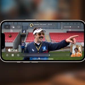 A smartphone displays a scene from the series "Ted Lasso" on Apple TV+, as a man with a mustache points and smiles. Thanks to Apple TV SharePlay, a small video call window appears in the corner, letting friends watch together. The Apple TV+ logo is visible.