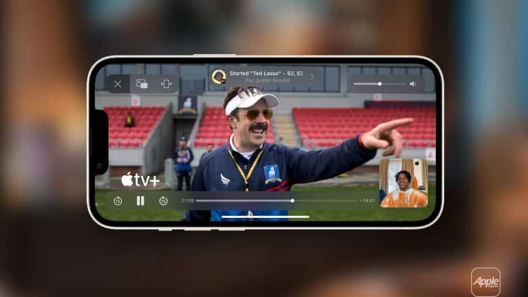 A smartphone displays a scene from the series "Ted Lasso" on Apple TV+, as a man with a mustache points and smiles. Thanks to Apple TV SharePlay, a small video call window appears in the corner, letting friends watch together. The Apple TV+ logo is visible.