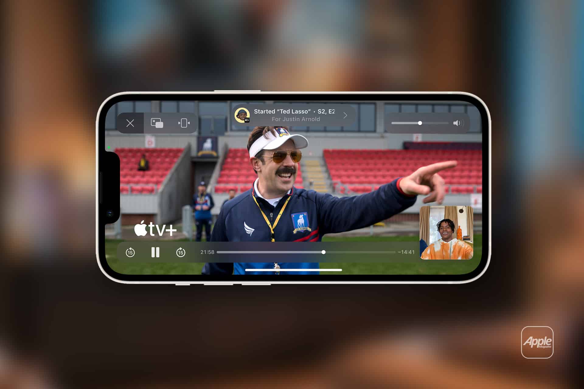 A smartphone displays a scene from the series "Ted Lasso" on Apple TV+, as a man with a mustache points and smiles. Thanks to Apple TV SharePlay, a small video call window appears in the corner, letting friends watch together. The Apple TV+ logo is visible.