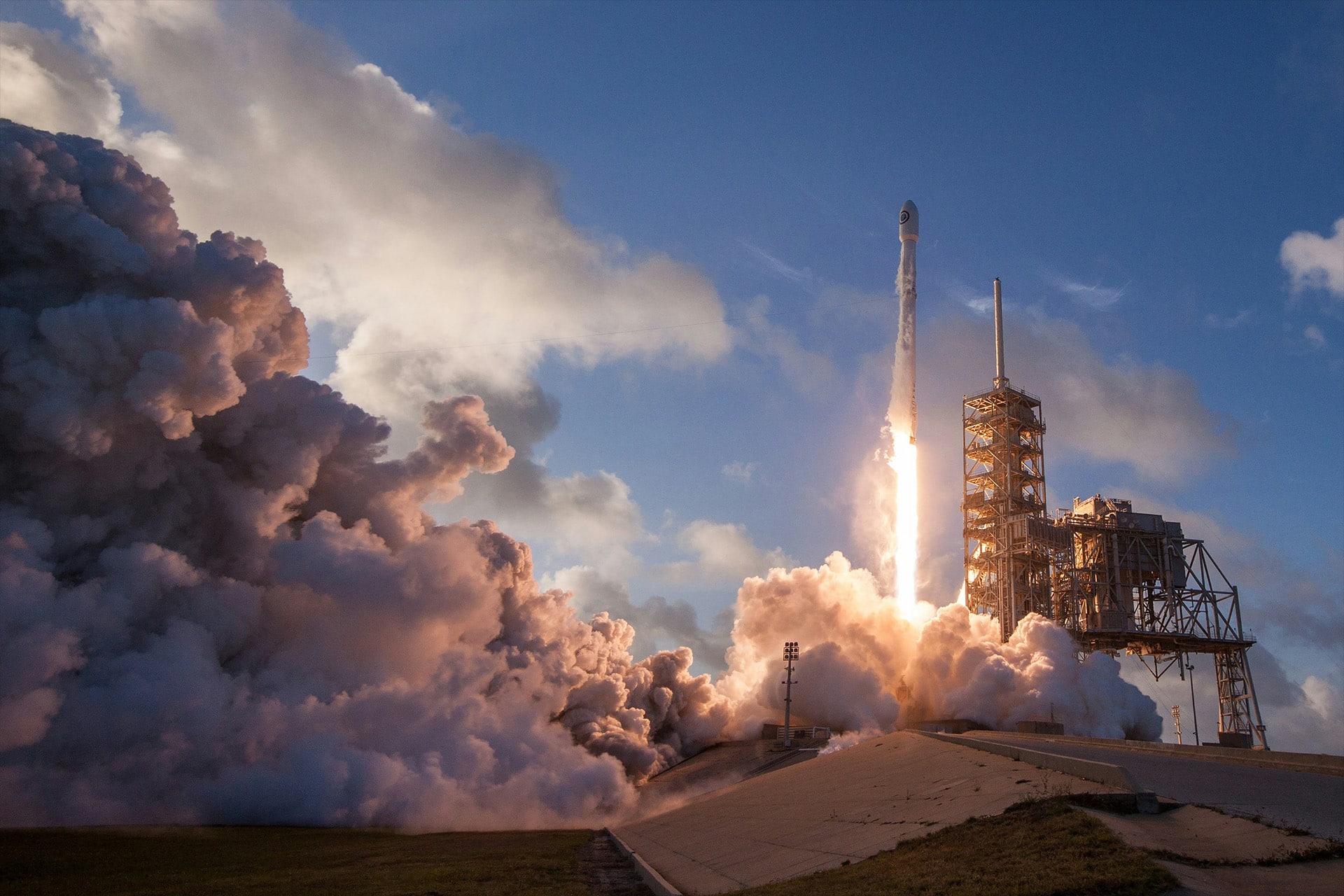 A SpaceX rocket launches into the sky from a launch pad, surrounded by thick clouds of smoke and flames, as part of the SpaceX and Apple satellite strategy, with a partly cloudy blue sky in the background.