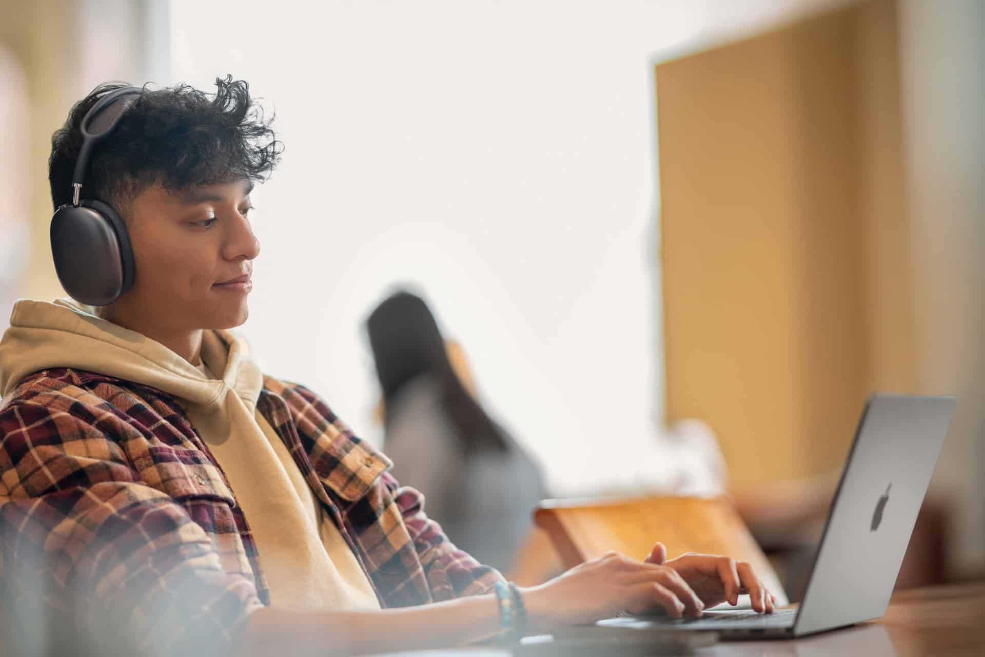 A young person wearing headphones and a plaid shirt over a hoodie uses a laptop at a desk in a bright, modern setting—possibly working on an Apple Developer Academy project. The background is softly blurred, showing another person sitting further away.