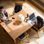 Two people sit across from each other at a wooden table, working on laptops. The table holds Xcode notebooks, headphones, and water bottles, with sunlight streaming in—a scene straight out of the Apple Developer Academy.