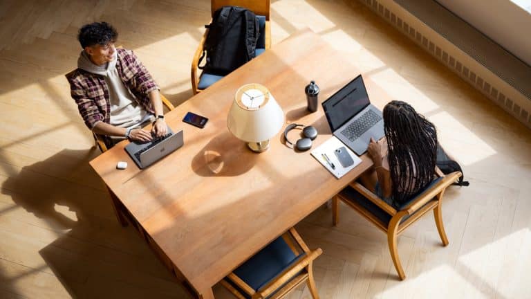 Two people sit across from each other at a wooden table, working on laptops. The table holds Xcode notebooks, headphones, and water bottles, with sunlight streaming in—a scene straight out of the Apple Developer Academy.