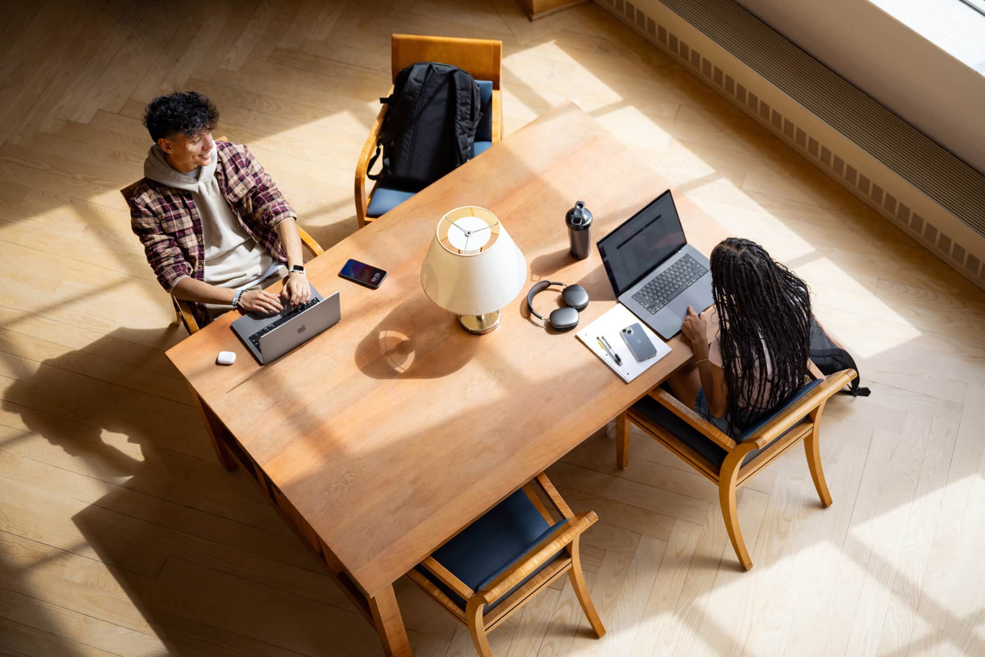 Two people sit across from each other at a wooden table, working on laptops. The table holds Xcode notebooks, headphones, and water bottles, with sunlight streaming in—a scene straight out of the Apple Developer Academy.