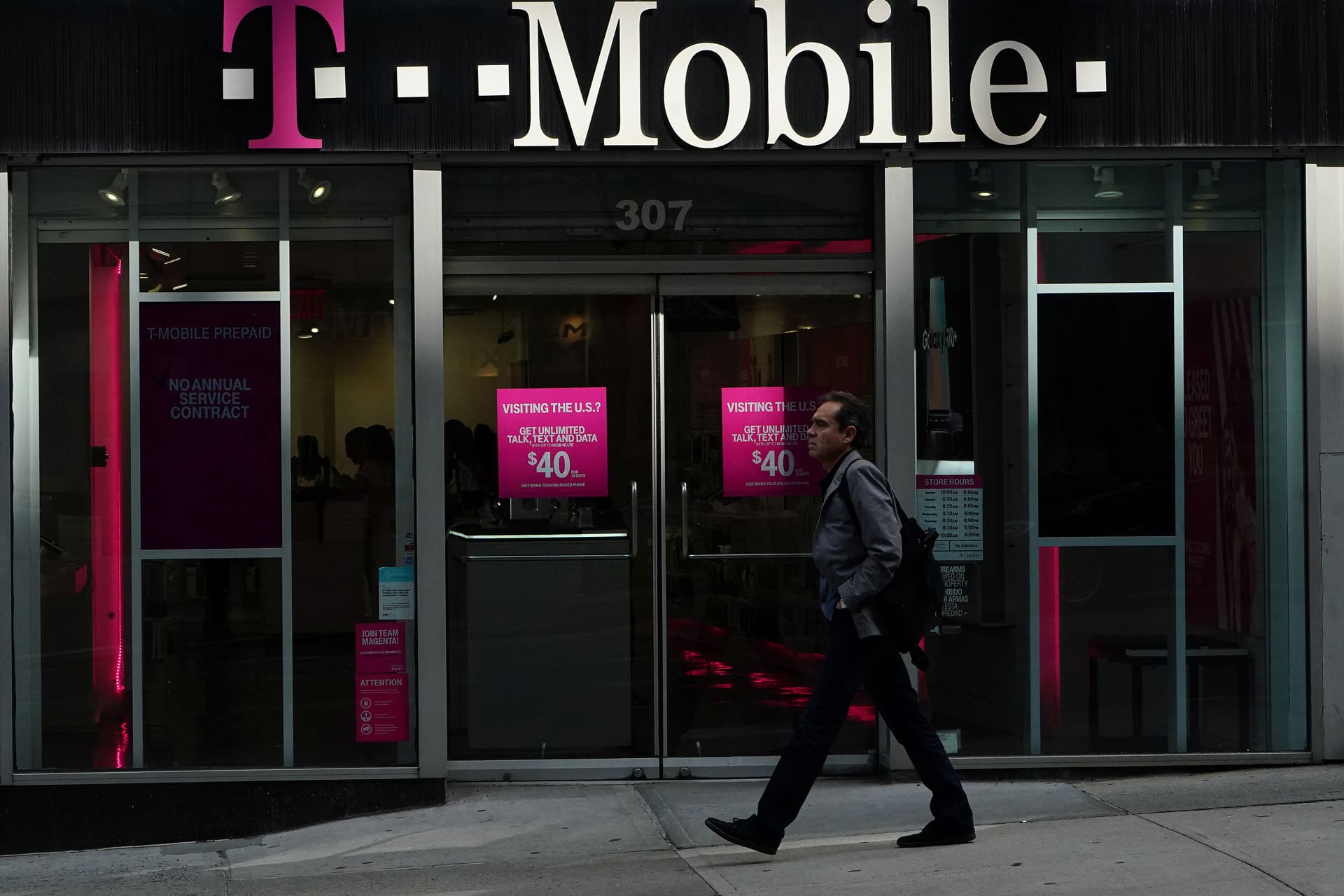 A person walks past a T-Mobile store with large glass doors and windows, displaying bright pink signs advertising $40 phone plans and iPhone satellite features. The T-Mobile logo is prominently visible above the entrance.