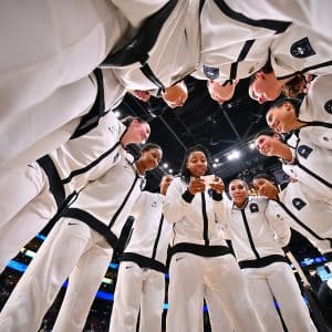 A group of basketball players in white tracksuits stand in a circle, smiling and listening, channeling the unity and spirit seen in The Dynasty: UConn Huskies before a big game on an indoor court.