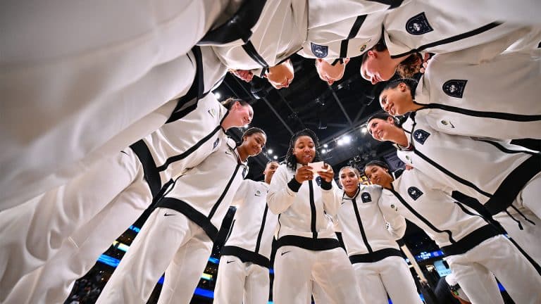 A group of basketball players in white tracksuits stand in a circle, smiling and listening, channeling the unity and spirit seen in The Dynasty: UConn Huskies before a big game on an indoor court.