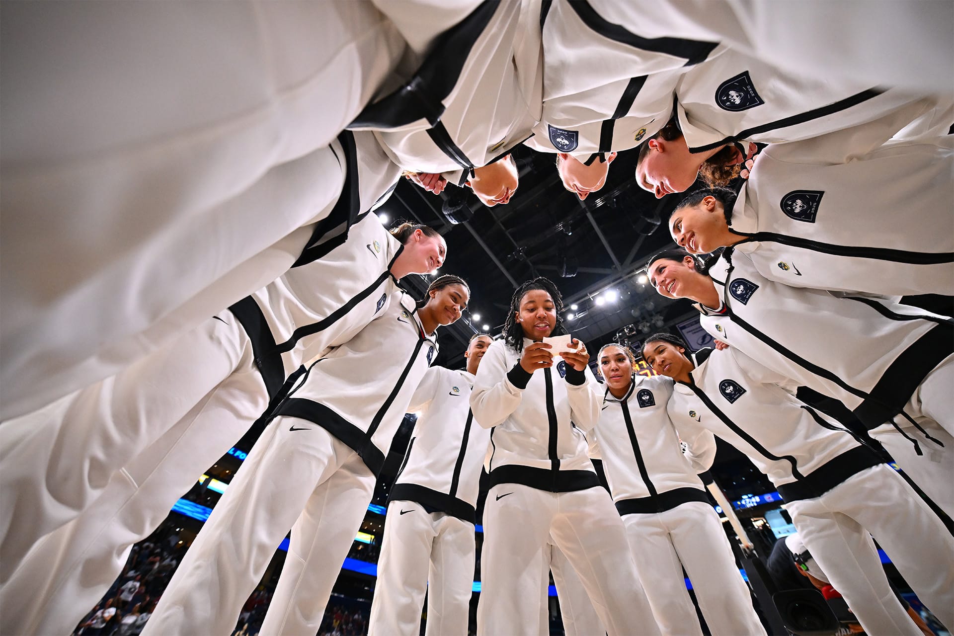 A group of basketball players in white tracksuits stand in a circle, smiling and listening, channeling the unity and spirit seen in The Dynasty: UConn Huskies before a big game on an indoor court.