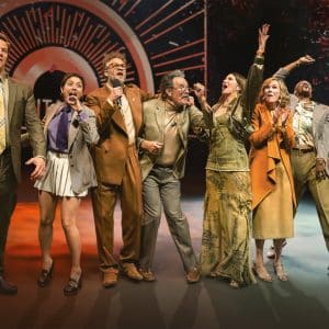 Eight people stand in a row on stage, excitedly smiling and cheering with arms raised, dressed in colorful and eclectic outfits, celebrating The Studio awards before a dramatic, illuminated backdrop.