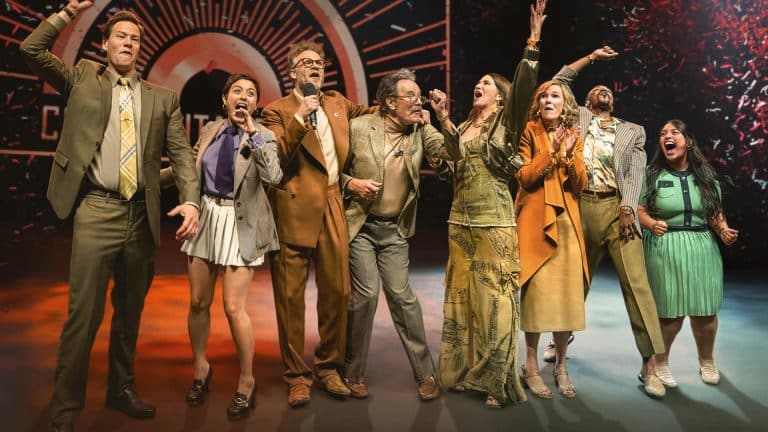Eight people stand in a row on stage, excitedly smiling and cheering with arms raised, dressed in colorful and eclectic outfits, celebrating The Studio awards before a dramatic, illuminated backdrop.