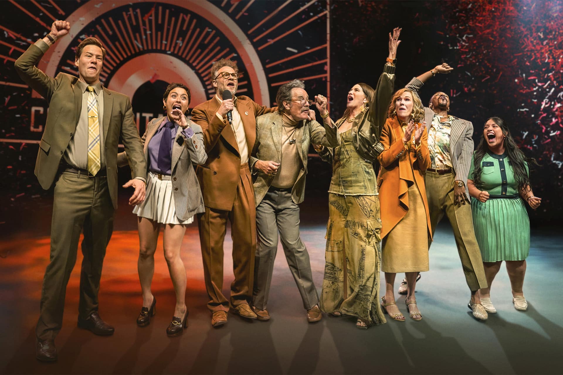 Eight people stand in a row on stage, excitedly smiling and cheering with arms raised, dressed in colorful and eclectic outfits, celebrating The Studio awards before a dramatic, illuminated backdrop.
