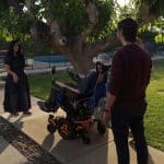 Three people interact outdoors near a tree—one uses a Vision Pro wheelchair, another stands nearby, and a third in a long dress holds an object on the walkway. The scene is sunny with green grass.
