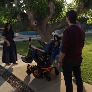 Three people interact outdoors near a tree—one uses a Vision Pro wheelchair, another stands nearby, and a third in a long dress holds an object on the walkway. The scene is sunny with green grass.