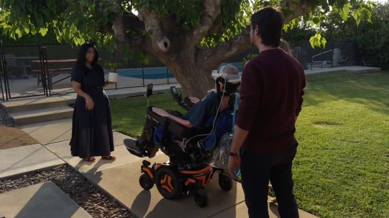 Three people interact outdoors near a tree—one uses a Vision Pro wheelchair, another stands nearby, and a third in a long dress holds an object on the walkway. The scene is sunny with green grass.