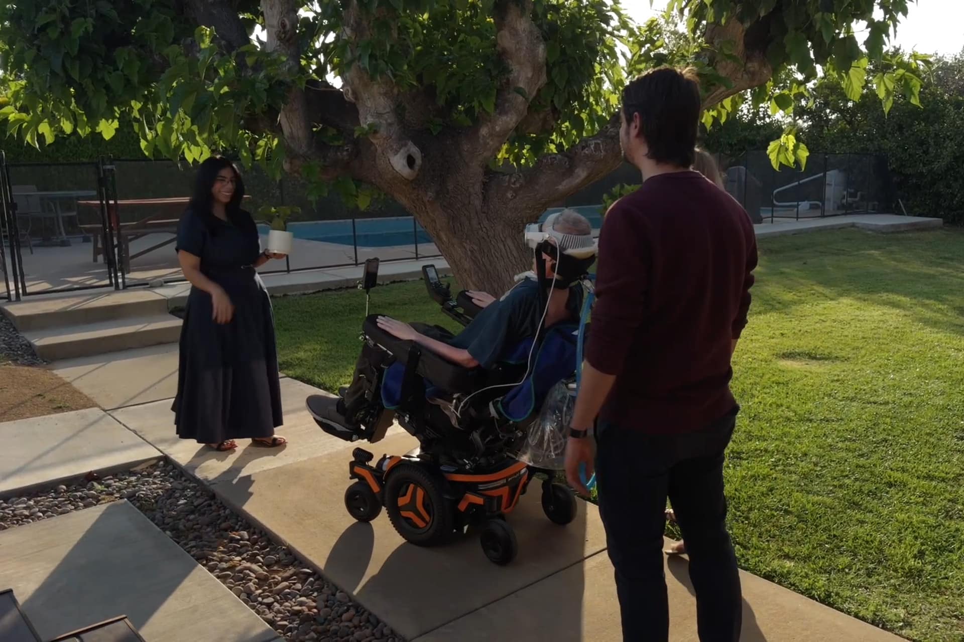 Three people interact outdoors near a tree—one uses a Vision Pro wheelchair, another stands nearby, and a third in a long dress holds an object on the walkway. The scene is sunny with green grass.