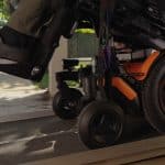 Close-up of an orange Vision Pro wheelchair as it moves up a ramp, with a person's foot resting on the footrest. The background shows a sunny outdoor area filled with greenery and pavement.
