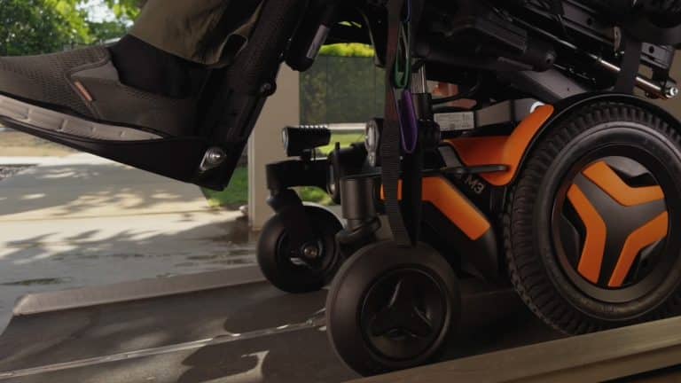 Close-up of an orange Vision Pro wheelchair as it moves up a ramp, with a person's foot resting on the footrest. The background shows a sunny outdoor area filled with greenery and pavement.