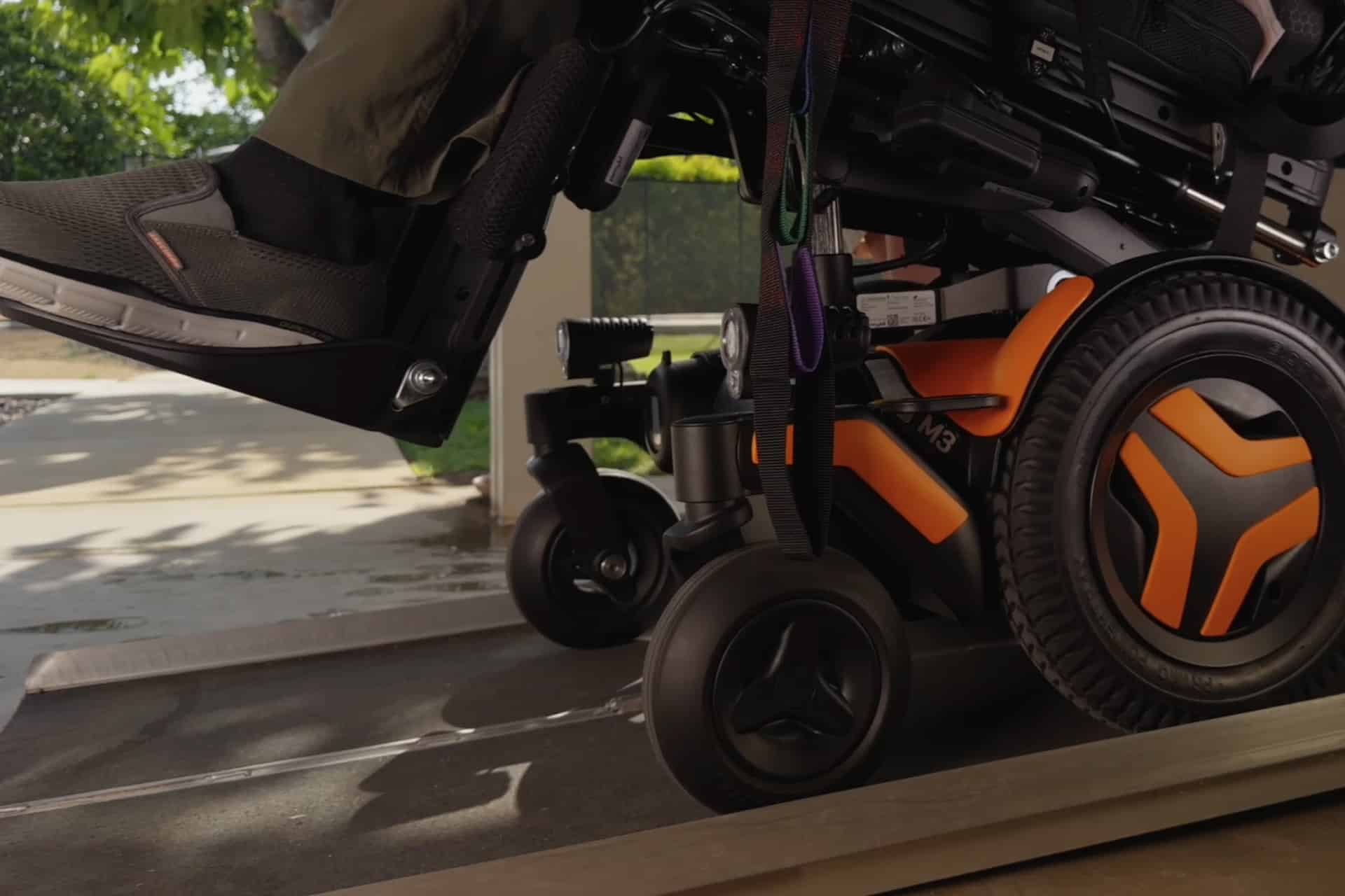 Close-up of an orange Vision Pro wheelchair as it moves up a ramp, with a person's foot resting on the footrest. The background shows a sunny outdoor area filled with greenery and pavement.
